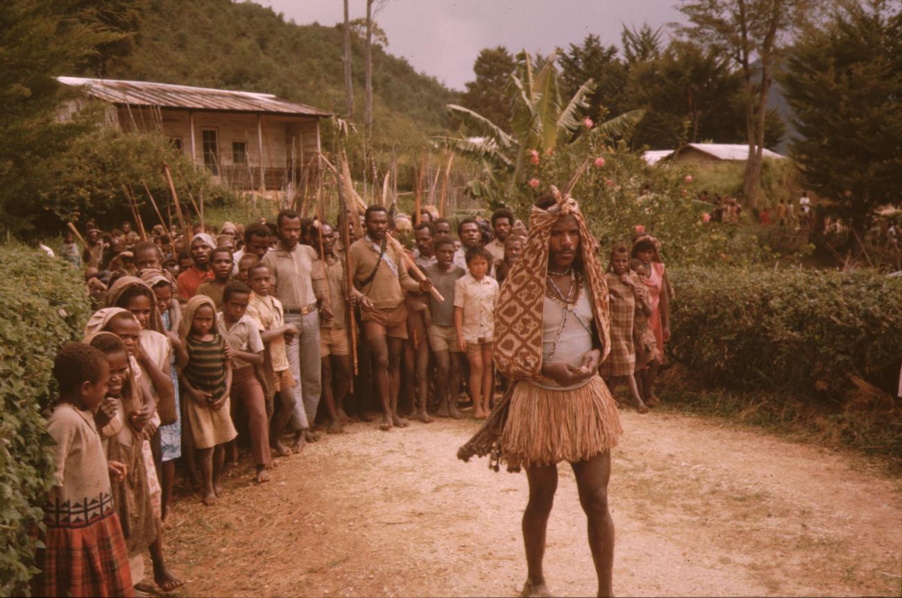 BD/132/128 - 
mannen en kinderen met speren achter een man in traditionele kleding
