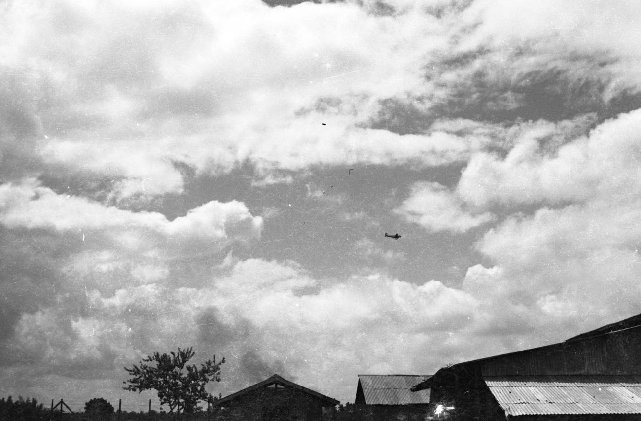 BD/133/1108 - 
Overkomend vliegtuig boven golfplaten daken
