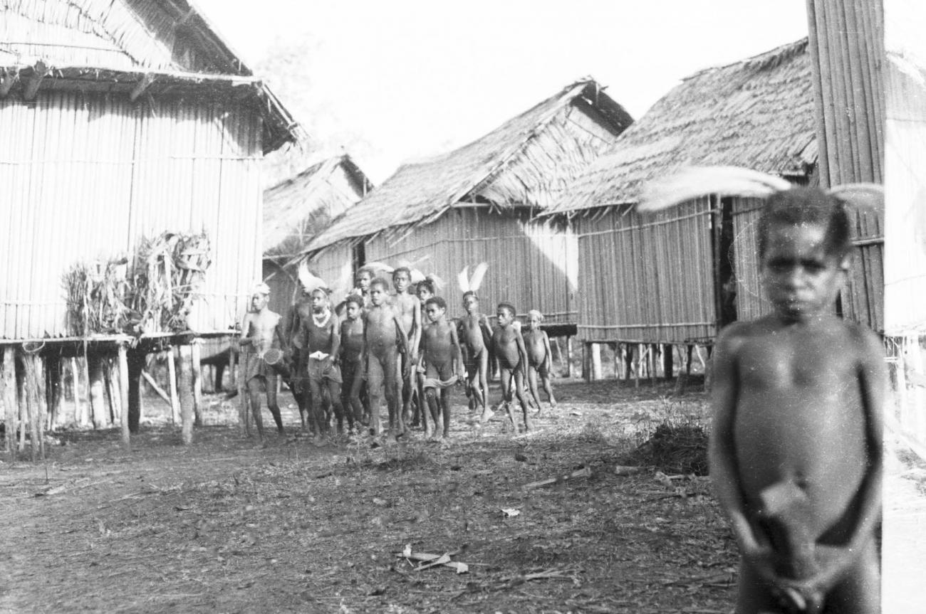 BD/133/448 -
Tocht Merauke-Kepi-Cookrivier vv: Portret van een jongen met paradijsvogelveren als hoofdsieraad en bamboekoker in zijn handen, met dorpsbewoners op achtergrond