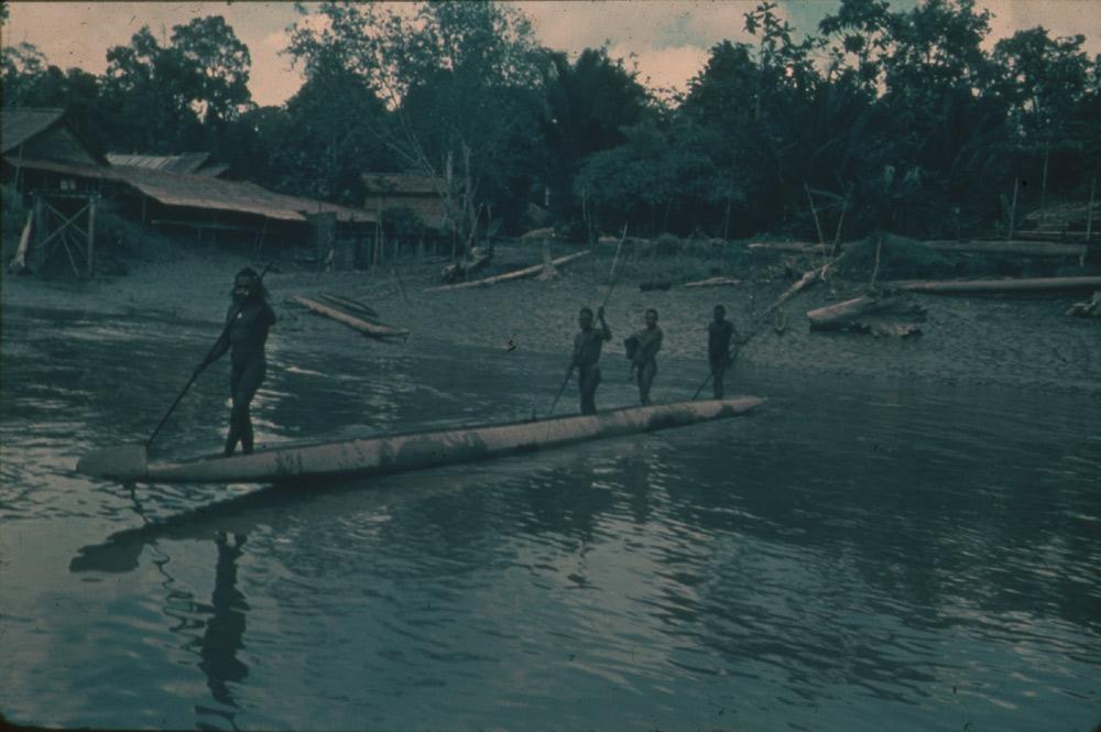 BD/30/3 - 
Asmatters in een prauw op de rivier voor een nederzetting met paalwoningen
