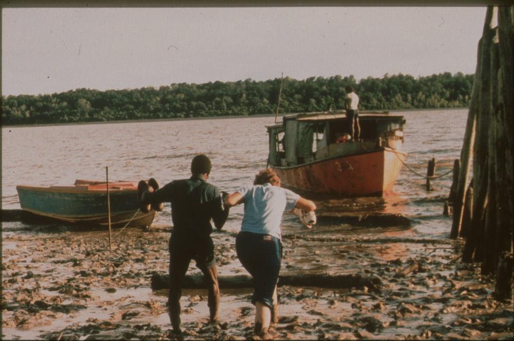 BD/30/64 - 
Asmatman helpt blanke vrouw door modder naar boot op rivier
