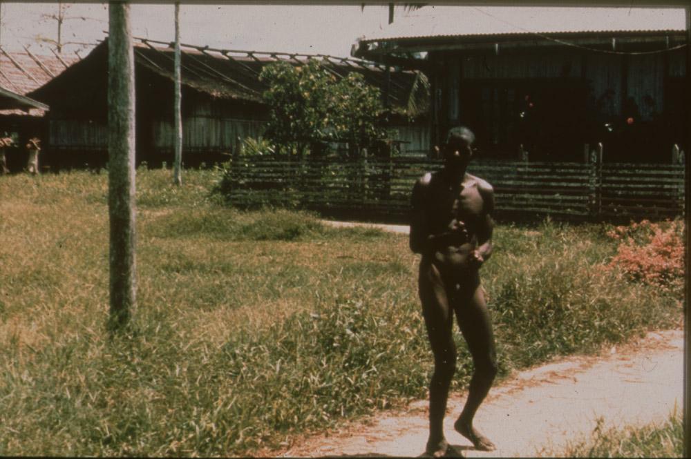 BD/30/93 - 
Asmatman met stok in hand staat op pad in dorp
