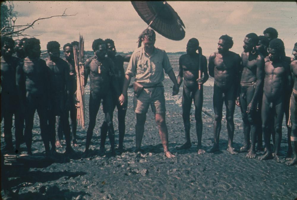 BD/30/99 -
Bestuursambtenaar staat onder parasol met Asmatmannen op modderig strand